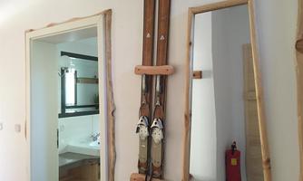 A modern ski hut with skis mounted on the wall and a large mirror. In the background, a bathroom with a sink and a illuminated mirror can be seen.