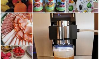 A varied breakfast with fresh fruit, cold cuts, and various drinks. In the center is a coffee machine with a glass of coffee.