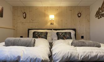 A cozy bedroom with two beds and modern bedcovers. The walls are adorned with wooden panels and there is atmospheric lighting.