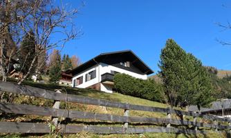 A modern house stands on a hill, surrounded by trees and a wooden privacy fence. The sky is clear and blue, creating a peaceful atmosphere.
