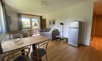 A bright living space with a sofa and a dining table. Large windows provide a view of nature and a modern kitchen unit with a refrigerator.