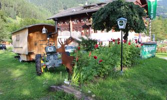 A traditional wooden trailer stands next to an old tractor in a picturesque outdoor area. Blooming flowers and a surrounding mountain landscape give the scene a rustic charm.