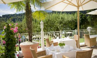 A cozy outdoor area of a restaurant with white tables and chairs. Palms and flowers decorate the terrace, surrounded by a green landscape.