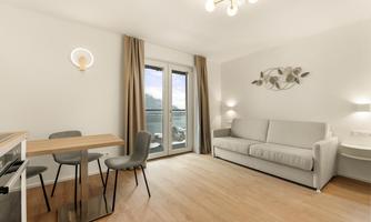 A bright living room with a gray sofa and a dining table. Large windows offer a view of the landscape.