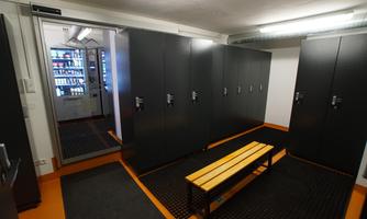 A modern changing room with black lockers and a wooden bench. A vending machine is visible in the background.