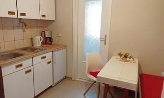 A small, bright kitchen with white cabinets and a table. There is a coffee machine and a refrigerator, as well as red seat cushions on the chairs.