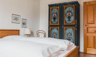 A cozy bedroom with a double bed and a colorful wooden wardrobe. The walls are plain and the furniture is made of light wood.