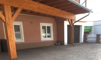 A covered area with wooden supports and paved floor. In the background, windows and a garage are visible.