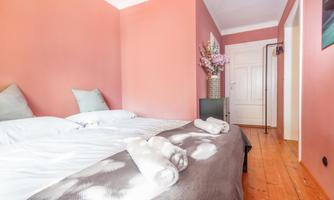 A modern bedroom with pink walls and a wooden floor. There are two beds, some cushions, and a vase with flowers.