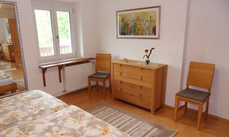 A bright room with a double bed and wooden furniture. There are two chairs, a dresser, and a large window.
