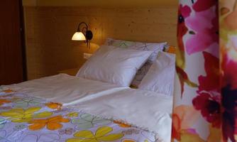 A cozy bedroom with a large bed and colorful floral bedding. The walls are in warm shades of yellow, and a bedside lamp emits soft light.