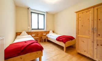 A simple bedroom with two single beds and wooden furniture. There is a window and a wooden chest of drawers.
