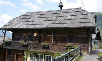 A traditional wooden house with a steep roof and a balcony. It is surrounded by green mountains and has a rustic, picturesque ambiance.