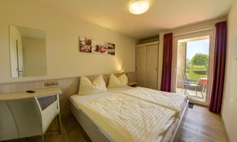 A bright room with a double bed and a modern desk. Large windows lead to a terrace with a view of greenery.