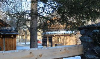 A snowy landscape with wooden buildings and tall trees. In the background, more wooden houses can be seen.