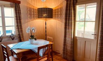 A cozy dining area with a wooden table and chairs. An orchid sits on the table, and a lamp on the wall provides warm light.