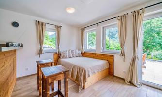 A cozy bedroom with a bed, two small tables, and large windows. The walls are light and there is a view of greenery.