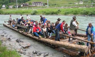 A group of people is sitting on a large raft in the river. In the background, green hills and buildings can be seen.