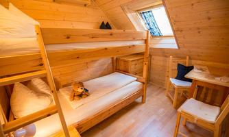 A cozy room with a bunk bed and a wooden single bed. The room is bright and friendly, with a small table and a window opening.