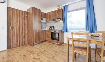 A modern kitchen with wooden furniture and an integrated cooking area. A dining table with chairs is next to the window, allowing daylight to come in.