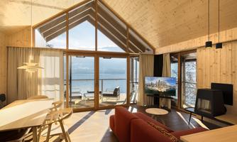 A modern living room with wooden paneling and large windows. View of the lake and a terrace with sun loungers.