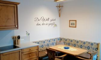 A cozy kitchen with wooden cabinets and a dining table. A saying hangs on the wall and there is a bench with a blue fabric cover.