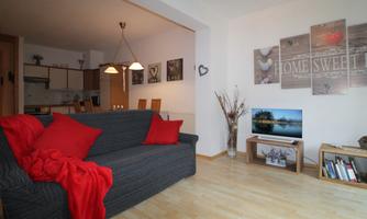 A cozy living room with a gray sofa and red cushions. Pictures hang on the wall and a television sits on a wooden shelf.