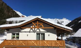 A cozy chalet in the snow with a wooden area and a beautiful viewpoint of the mountains. The clear blue sky creates a charming winter landscape.