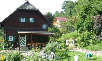 A pretty, traditional wooden house with a blooming garden. The surroundings are green and inviting, characterized by flowers and trees.