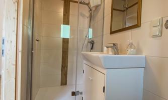A modern bathroom with a shower and a sink. The walls are tiled in light colors, and wooden details create a cozy atmosphere.