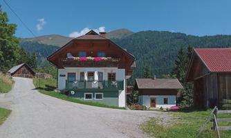 A charming farmhouse with balconies and colorful flowers. In the background, green mountains and a clear sky can be seen.