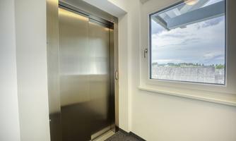 A modern elevator in a bright hallway. Through the window, one can see a cloudy landscape.
