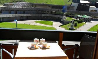 A balcony with a small table on which two cups and a few snacks are placed. In the background, a green landscape with mountains can be seen.
