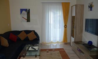 A cozy living room with a blue couch and colorful cushions. Bright curtains hang in front of the window, and the television is on a modern shelf.