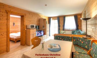 A cozy holiday apartment with wooden paneling and a table in the foreground. In the background, there is a bedroom and a living area with a TV and kitchenette.