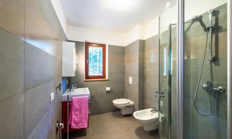 A modern bathroom with gray tiles and a large shower. The room includes a bidet and a washbasin with a mirror.