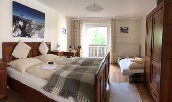 A cozy bedroom with two beds, a small table, and a large window. The wooden furniture and a soft blanket create a warm atmosphere.