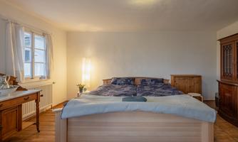A cozy bedroom with a large bed and wood-colored furniture. The room has large windows and a light source that creates a pleasant atmosphere.