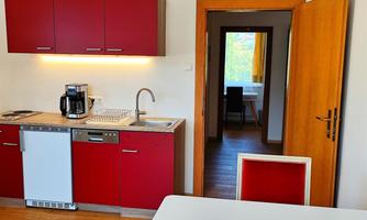 A modern kitchen with red cabinets and a wooden floor. In the background, an open room with a table and a window is visible.