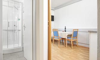 A bright room with a table and chairs as well as a shower in the background. The floor is made of wood and the walls are painted white.