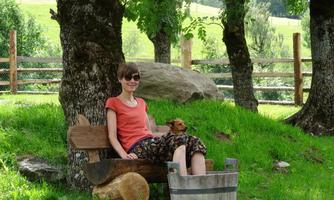 A woman is sitting on a wooden bench outdoors with a dog on her lap. In the background, trees and a meadow can be seen.