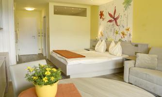 A bright room with a double bed and a cozy seating area. Colorful flower patterns can be seen on the wall, and a blooming pot sits on the table.