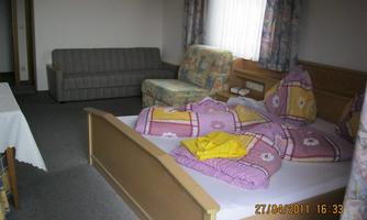 A simple room with a double bed, colorful bedspreads, and a sofa. There is also a small table and windows with curtains.