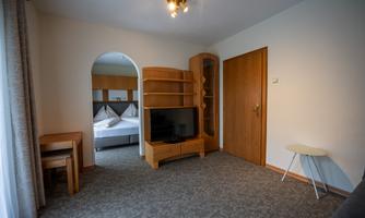 A simple room with a television, a table, and chairs. In the background, a bed is visible.