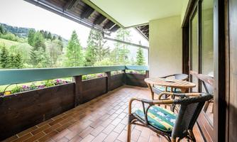 A cozy balcony with two chairs and a table. The view overlooks green trees and a picturesque landscape.