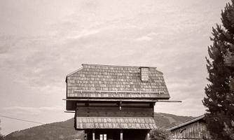 A traditional wooden house with a balcony and shingle roof. It stands on a gentle hill, surrounded by trees and meadows.