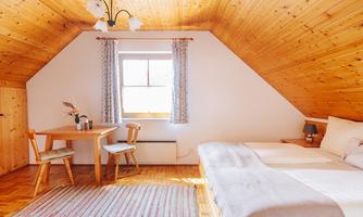 A cozy room with a wooden panelled ceiling and two windows. There is a double bed, a dining table with chairs, and a carpet on the floor.