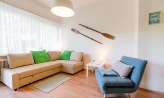 A bright living room with a beige sofa and green cushions. Oars hang on the wall, and there is a blue armchair next to a small table.