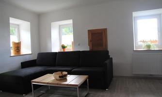 A modern living room with a black sofa and a wooden table. 

Large windows let in plenty of daylight, and the walls are painted in neutral colors.