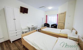 A simple room with two single beds, a table, and two chairs. There is a television, a wardrobe, and large windows with daylight.